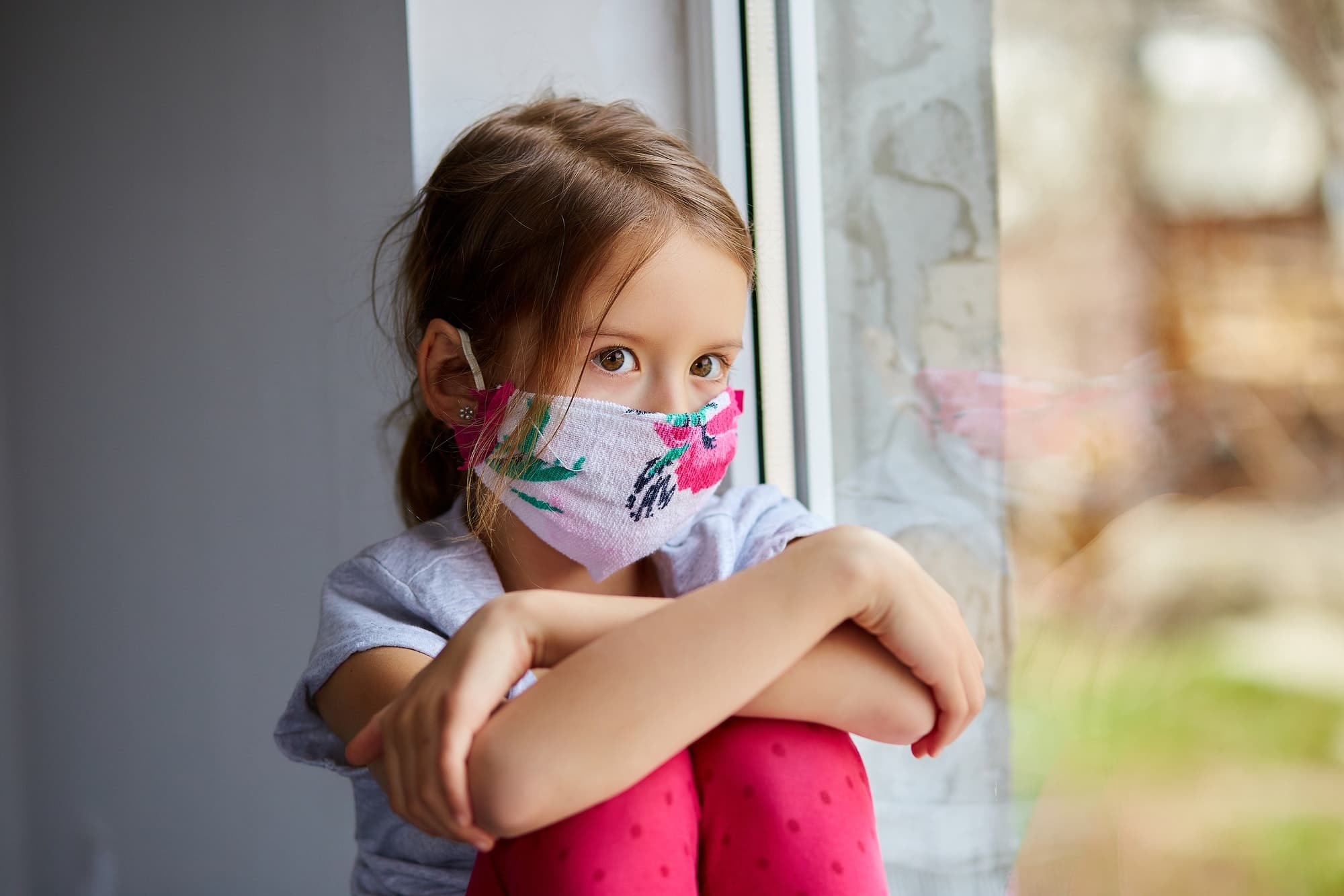 little girl child in mask sits on windows coronavirus quarantine